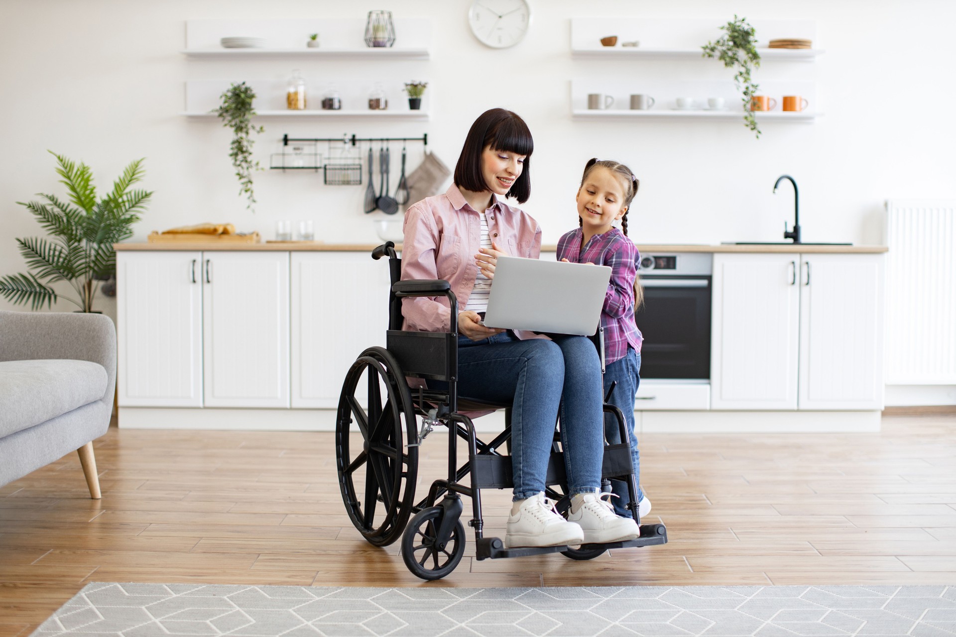 Mother in wheelchair using laptop with daughter in kitchen